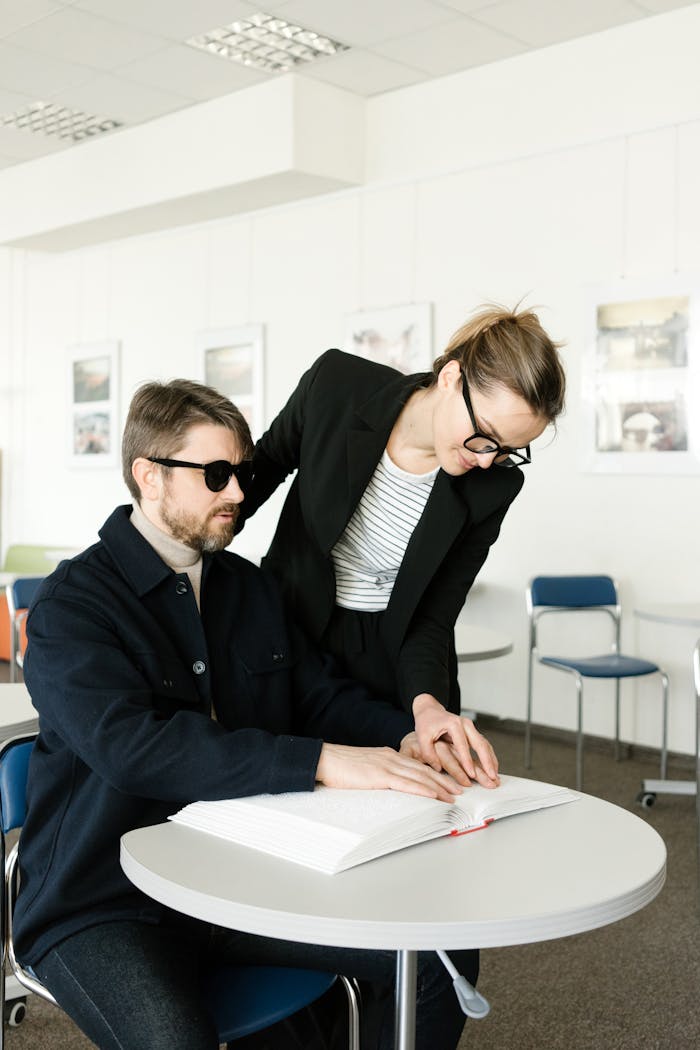A woman helps a blind man read braille in a modern classroom setting.
